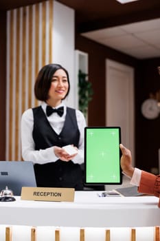 Woman holding tablet with greenscreen at front desk, receiving room access card from asian receptionist. Young person using isolated mockup display with chromakey at hotel reception.