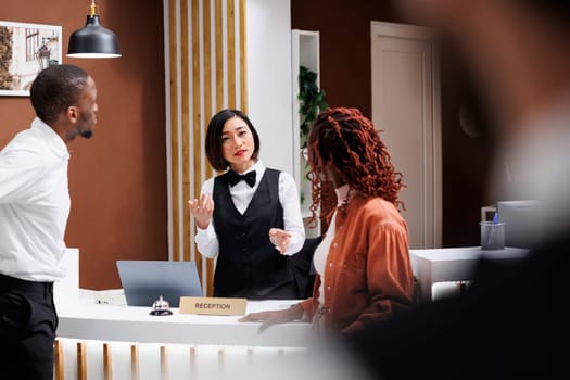 Worker talking to couple at reception desk in lobby, providing luxury service to guests. Asian receptionist welcoming people at modern expensive hotel, doing registration at entrance.