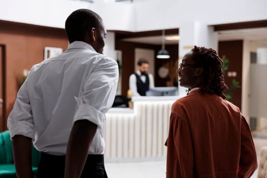 Guests arriving at hotel reception in lobby, entering lounge area and talking to receptionist about room reservation. Young couple asking front desk staff about accommodation and service.