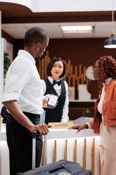 Young man paying for booking with mobile phone nfc, talking to receptionist about luxury resort services. Tourists making payment at pos terminal, filling in registration forms.