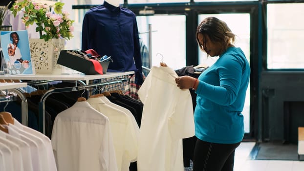 African american customer shopping for white shirt, checking items fabric in modern boutique. Shopaholic woman buying new fashion collection, looking at casual wear in clothing store