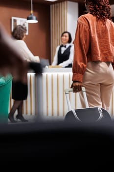 Tourist waiting to register at hotel, carrying bags in lobby area with reception counter. Woman with luggage preparing to check in and ask about room accommodation at front desk.