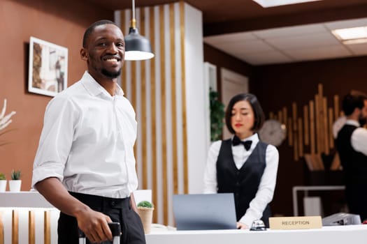 Male tourist sitting at front desk to register and fill in check in forms, guest smiling and waiting for hotel service. Young man standing near reception counter in lobby, luxury resort.