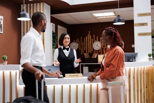 Asian woman helping guests to check in at reception desk, using records and registration forms for accommodation. Receptionist registering and welcoming people at hotel counter.