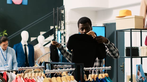 Stylish client shopping for fashionable clothes, analyzing merchandise material in clothing store. African american man looking at hangers with new fashion collection, buying basic outfits