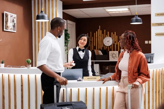 Receptionist giving room key card to couple arriving at hotel and doing check in registration, helping with accommodation. Guests preparing to have access to room after registering.