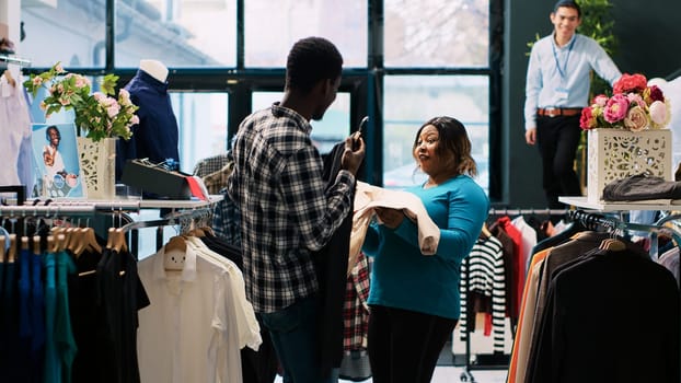 Cheerful couple playing with stylish merchandise, fooling with trendy clothes in modern boutique. African american customers shopping for fashionable items for new wardrobe. Fashion concept