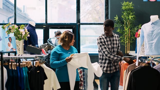 African american couple looking at fashionable clothes, checking items material in clothing store. Cheerful clients enjoying shopping session, buying stylish merchandise and accessories in modern boutique