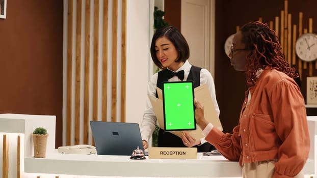 Weary tourist holding chroma key green screen mock up tablet at hotel reception desk after long flight, waiting for room payment to be processed. Guest in resort foyer doing check in