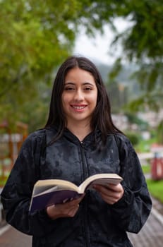 young and pretty latina walking in a park with a book on a rainy day looking at the camera. book day. High quality photo