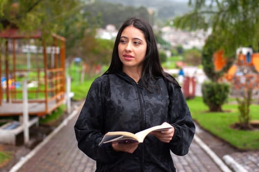 attractive young latina walking in a park with a book in her hand looking to the side on a morning walk. book day. High quality photo