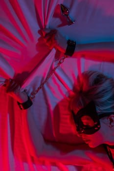 Sexy blonde woman in a leather mask and handcuffs and a collar lies in the bedroom in a red-blue neon light. Role-playing games