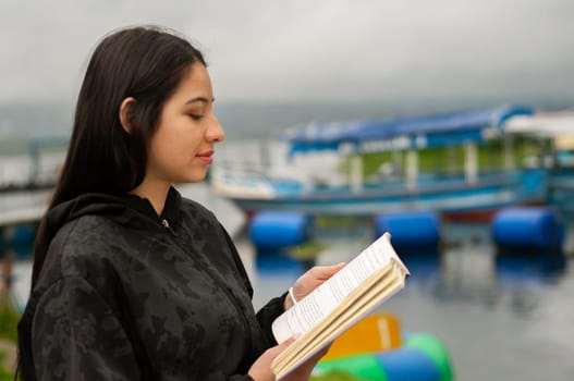 beautiful venezuelan woman in profile reading a novel on paper at the edge of a lake with blue boats. book day. High quality photo