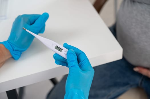 Pregnant woman with fever at doctor's appointment. Therapist holds an electronic thermometer with a temperature of 37.5