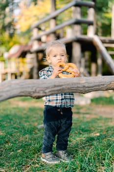 Little girl nibbles on a big bagel holding it with both hands in the park near the driftwood. High quality photo