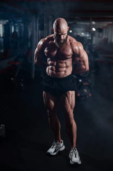 Muscular bald man posing in shorts. Bodybuilder showing off his shape in the gym