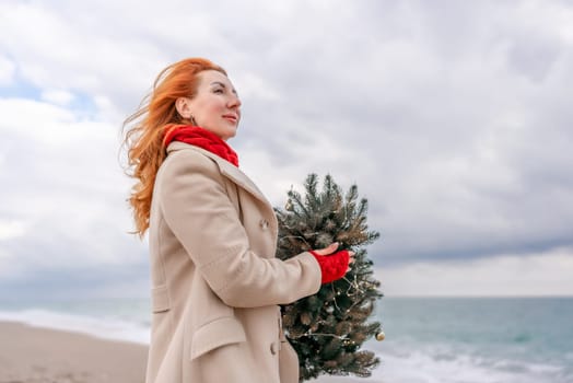 Redhead woman Christmas tree sea. Christmas portrait of a happy redhead woman walking along the beach and holding a Christmas tree in her hands. Dressed in a light coat, white suit and red mittens