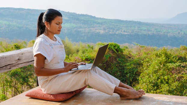 A young Asian businesswoman works at the computer in a cafe on the rock. Young girl downshifter working at a laptop at sunset or sunrise on the top of the mountain