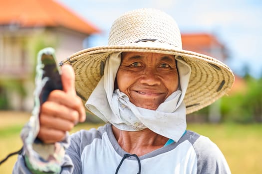 Emotional portrait of an Asian farmer wearing a straw hat and holding a sickle knife and cutting rice stalks in the field on a hot day. Bali, Indonesia - 12.03.2022