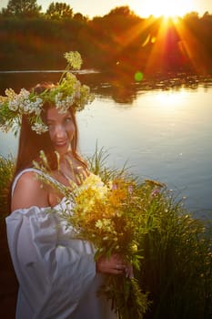 Adult mature brunette woman in a white dress, sundress and a wreath of flowers in summer by the water of river or lake in evening at sunset. Celebration of the Slavic pagan holiday of Ivan Kupala