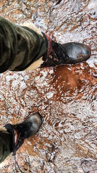 Feet of Hunter or fisherman in big warm boots on winter day on dirty snow. Top view. Fisherman on the river, lake, reservoir on spring day with melting ice. Dangerous fishing in spring or autumn day