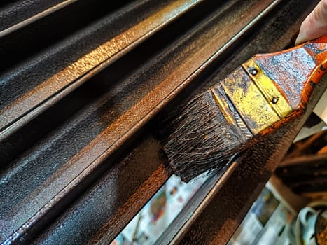 A brush in woman's hand paints iron metal bars with brown paint. Work and worker