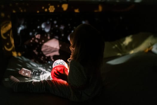Little girl sits on the bed with a night-light projector in her hands and looks at the pictures on the walls. High quality photo