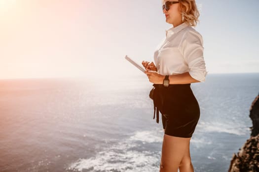 Successful business woman in yellow hat working on laptop by the sea. Pretty lady typing on computer at summer day outdoors. Freelance, travel and holidays concept.