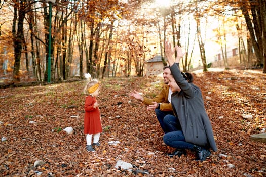 Little girl stands in front of mom and dad squatting with open arms in the forest. High quality photo