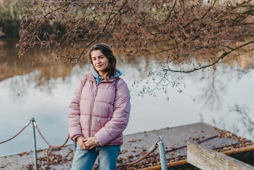 A young woman standing at the shore looking at the river in autumn sunny day. Street view, copy space for text, travel photo. Happy tourist woman on the bank of the river in autumn in warm clothes. Tourists enjoy their vacation, winter season. Romantic look and travel concept. A joyful mood in a Caucasian girl. Winter Wonderland: Enchanting Girl by the Riverside in Autumn.