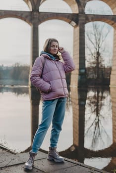 A young woman standing at the shore looking at the river in autumn sunny day. Street view, copy space for text, travel photo. Happy tourist woman on the bank of the river in autumn in warm clothes. Tourists enjoy their vacation, winter season. Romantic look and travel concept. A joyful mood in a Caucasian girl. Winter Wonderland: Enchanting Girl by the Riverside in Autumn.