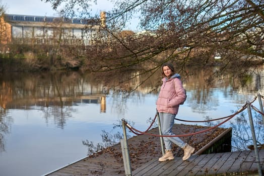 A young woman standing at the shore looking at the river in autumn sunny day. Street view, copy space for text, travel photo. Happy tourist woman on the bank of the river in autumn in warm clothes. Tourists enjoy their vacation, winter season. Romantic look and travel concept. A joyful mood in a Caucasian girl. Winter Wonderland: Enchanting Girl by the Riverside in Autumn.