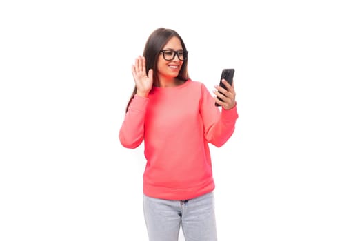 pretty young brunette caucasian woman with straight hair dressed in a pink sweatshirt and jeans chatting by video call on a smartphone on a white background with copy space.