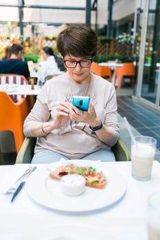 European mature woman sitting at table on terrace of restaurant drinking milk cocktail and chatting on mobile app.