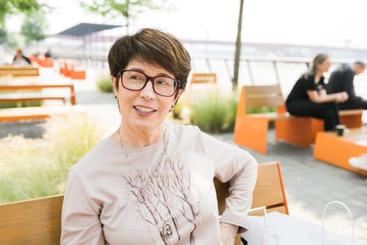 Outdoor portrait of beautiful and elegant middle age 55 - 60 year old woman wearing long sleeve top sitting on bench on the street. Copy space and empty place for text advertising.