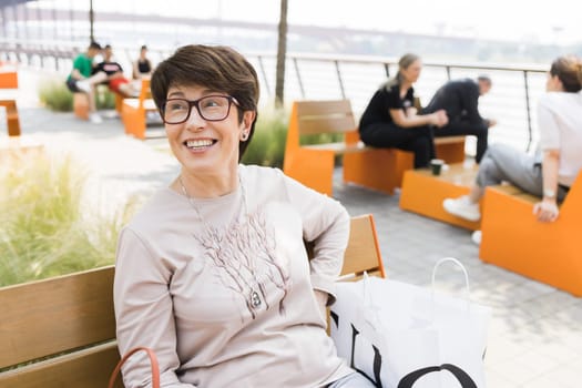 Outdoor portrait of beautiful and elegant middle age 55 - 60 year old woman wearing long sleeve top sitting on bench on the street. Copy space and empty place for text advertising.