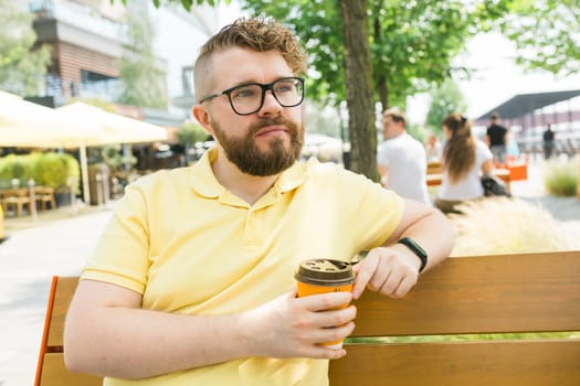 Millennial smiling student man in yellow polo t-shirt eyeglasses sitting on bench drink coffee have breakfast rest relax in sunshine spring green city park outdoors on nature. Urban leisure concept