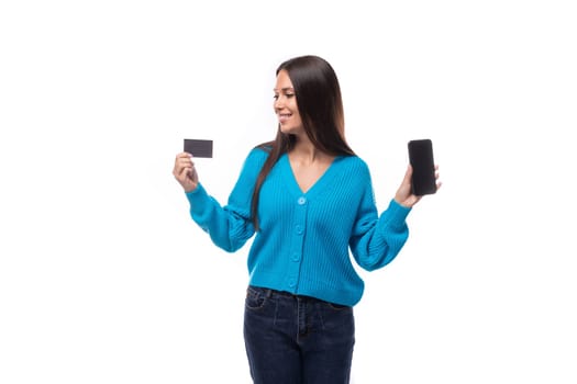 young beautiful european brunette woman dressed in a blue cardigan holds a bank card mockup and a smartphone.