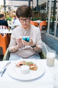 European mature woman sitting at table on terrace of restaurant drinking milk cocktail and chatting on mobile app.