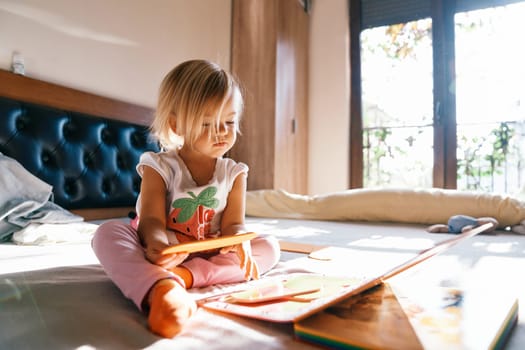 Little girl examines colorful puzzles, sitting on the bed in the room. High quality photo