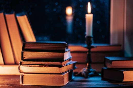 Stack of old paper books on wooden windowsill, bathed in soft candle glow light. Cozy ambience of fall, candle burning. Literature promotions or tranquil visual storytelling. Rainy autumn weather.