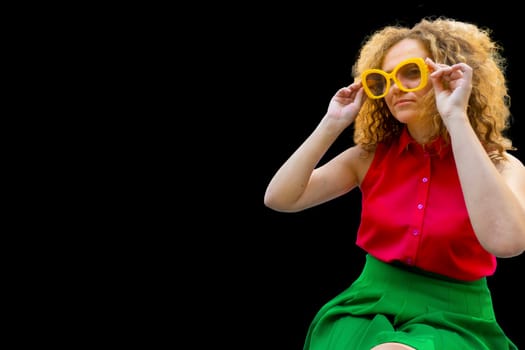 Young blonde woman with long curly hair in red and green fashion closes in sun glasses playful smile happy positive adjusts his glasses
