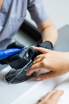 Female master using electric nail file while doing manicure for client in beauty salon.