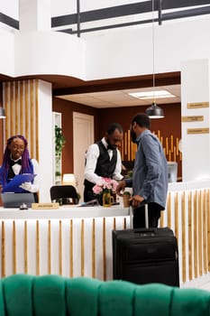 African American hotel front desk staff assist guests at reception, checking in tourists, requesting for reservation details during check-in. Tourist with luggage arriving at resort booking room