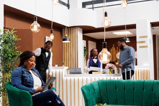 African American woman sitting in lobby with tablet using digital technology to check-in into hotel, tourist holding electronic device browsing internet while resting at resort foyer