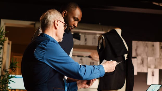 Couturier and pupil checking sketches in tailor workshop, learning to craft clothing line handmade. Fashion designer and apprentice discussing about models and fabric. Handheld shot.