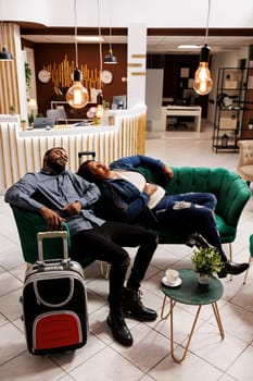 Tired couple sleeping in hotel lobby after long international flight, suffering from travel fatigue and jet lag. Exhausted tourists taking nap lying on sofa while waiting for check-in at resort