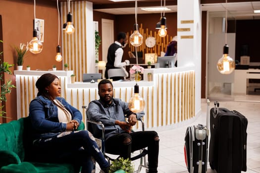 Young African American disabled man wheelchair user traveling with personal assistant, handicapped guy and his wife sitting in hotel lobby with luggage. Guest experience for people with disabilities