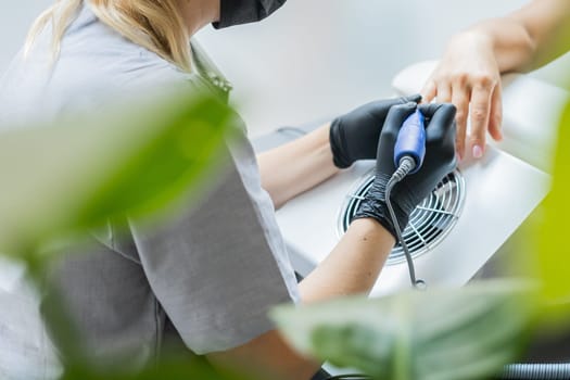 Female master using electric nail file while doing manicure for client in beauty salon.