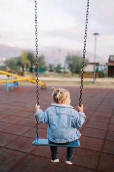 Little girl sits on a chain swing in the playground and looks away. Back view. High quality photo